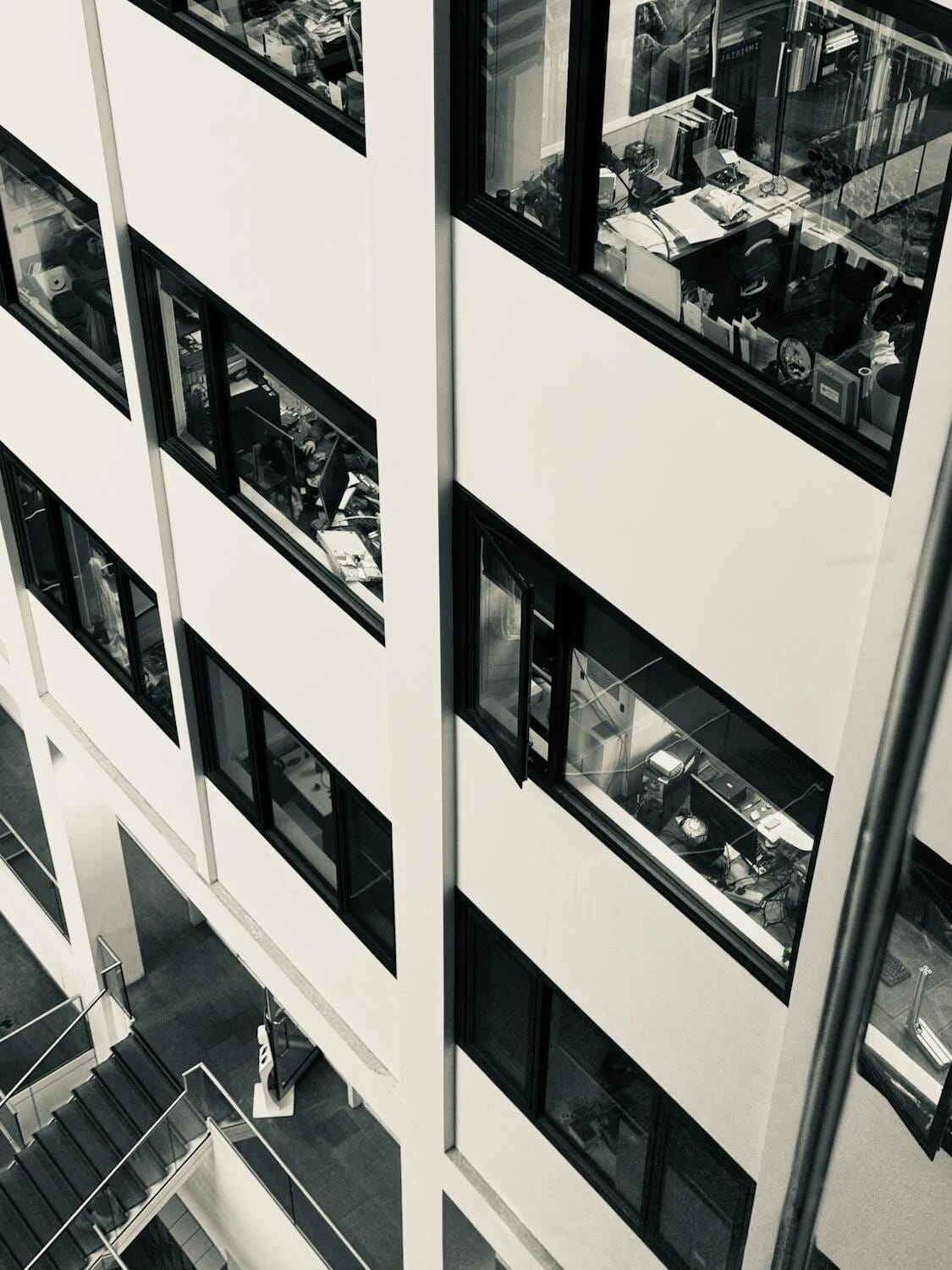 Photo en noir et blanc montrant plusieurs étages d'un immeuble de bureaux avec des bureaux et des espaces de travail encombrés visibles à travers de grandes fenêtres.