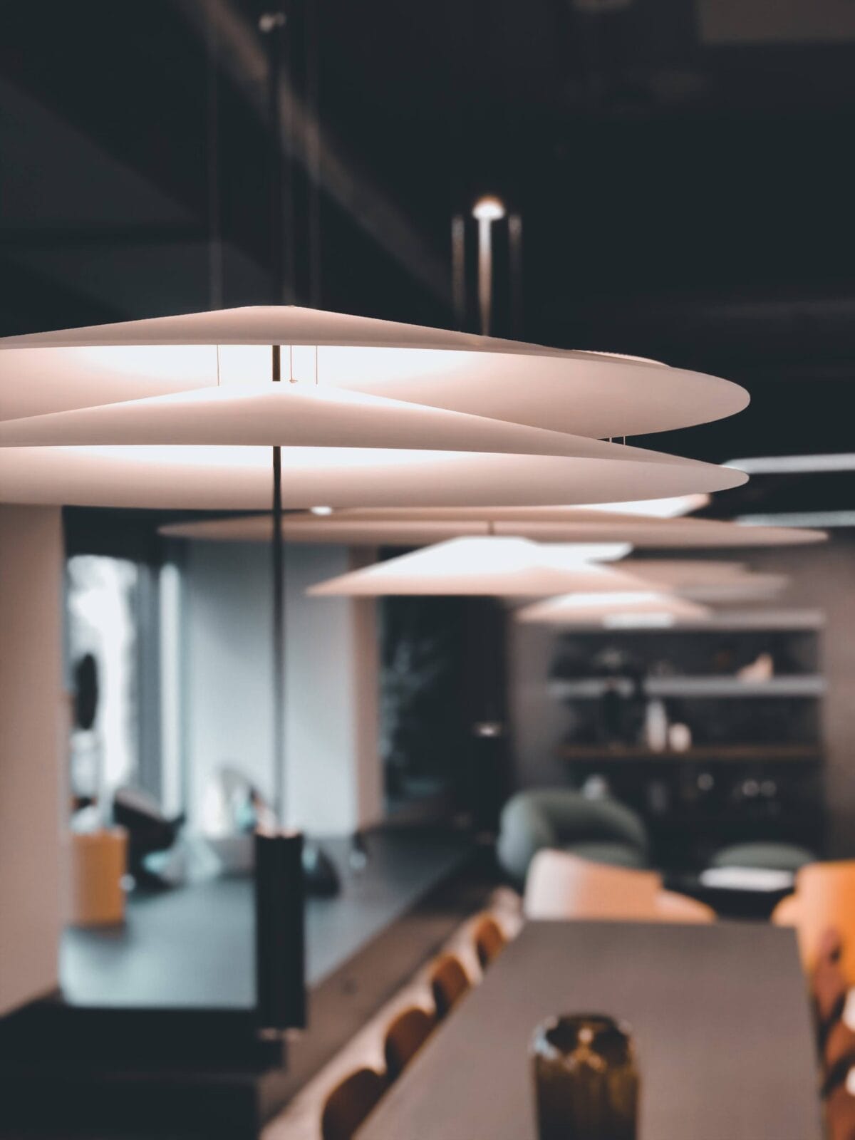 Un intérieur de bureau élégant avec des luminaires suspendus superposés épurés, une longue table avec des chaises, et un arrière-plan flou d'un espace salon moderne.