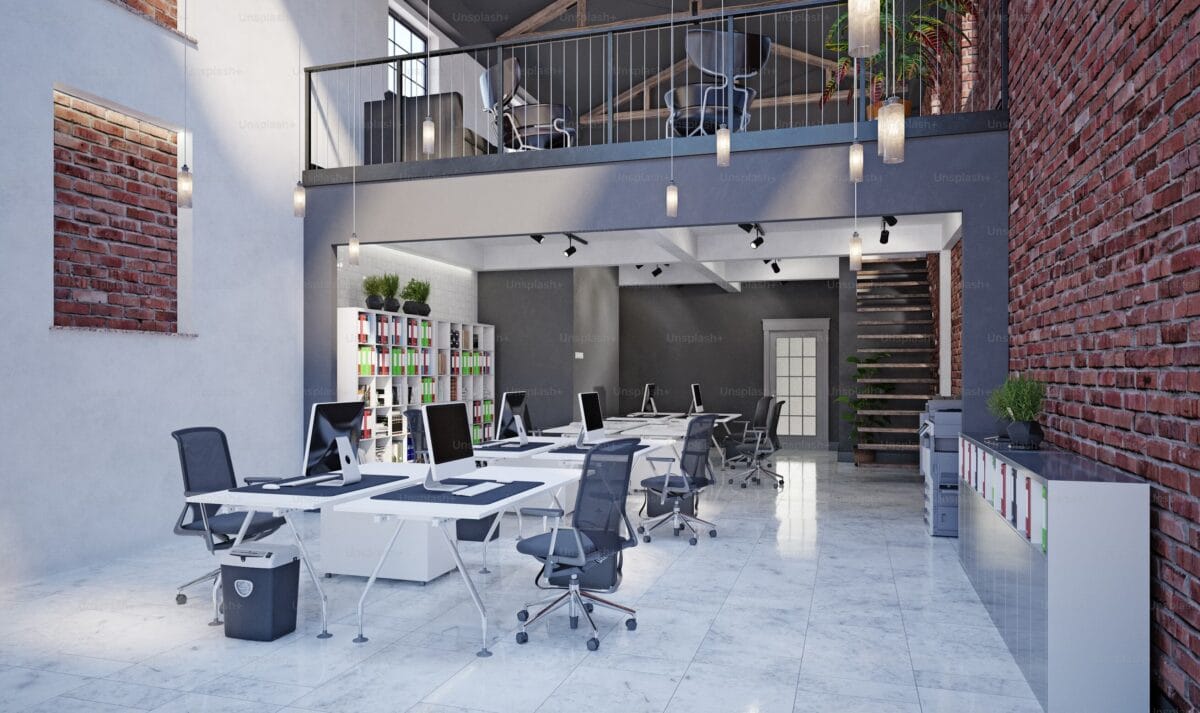 Bureau aménagé dans un loft avec murs en briques rouges, étage supérieur, chaises ergonomiques, étagères colorées, suspensions modernes et escalier en bois.
