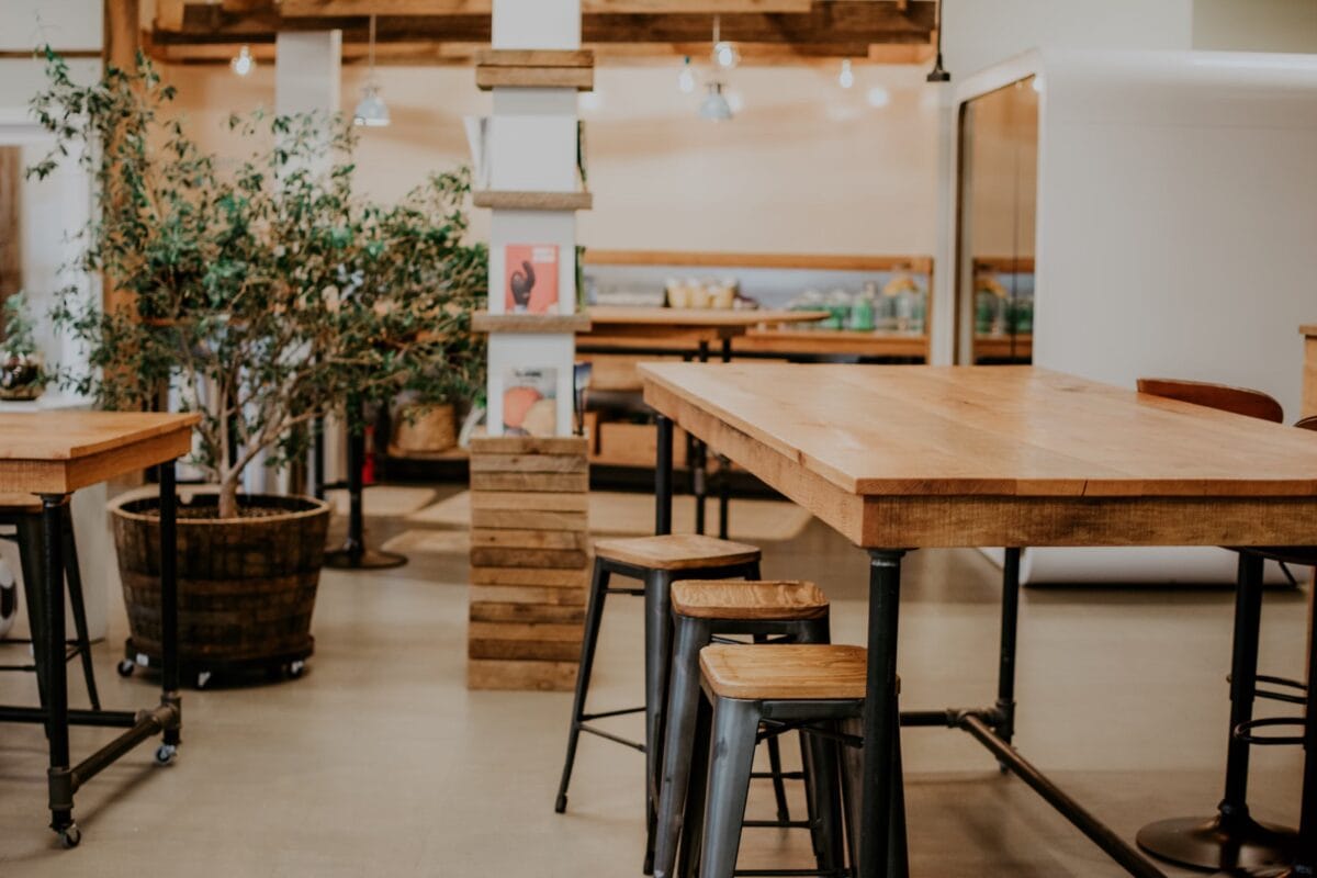 Espace de coworking cosy avec tables en bois, tabourets industriels, plantes et ambiance décontractée.